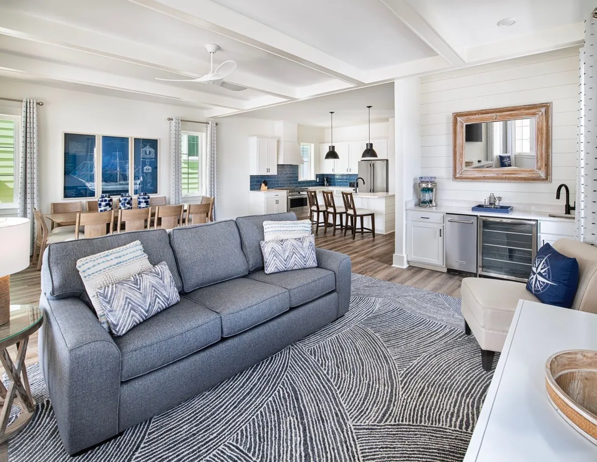 This image showcases a harmonious open-concept interior where the living area, kitchen, dining table, and wet bar are all visible. The gray sofa with patterned pillows creates a cozy social space, while the kitchen island with bar stools and pendant lights invites casual gatherings. The wet bar area, complete with a wine fridge and mirror, is seamlessly integrated for entertaining. Coastal décor elements and natural light enhance the relaxed, beach-inspired atmosphere.