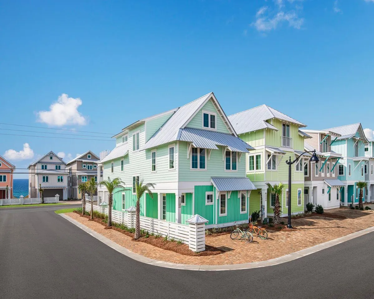 A view down a palm-lined street of colorful coastal-style houses with metal roofs, with the ocean visible at the end and bicycles parked outside. The scene evokes a cheerful, relaxing community vibe, inviting guests to explore the resort, stroll to the beach, or gather on balconies and patios.