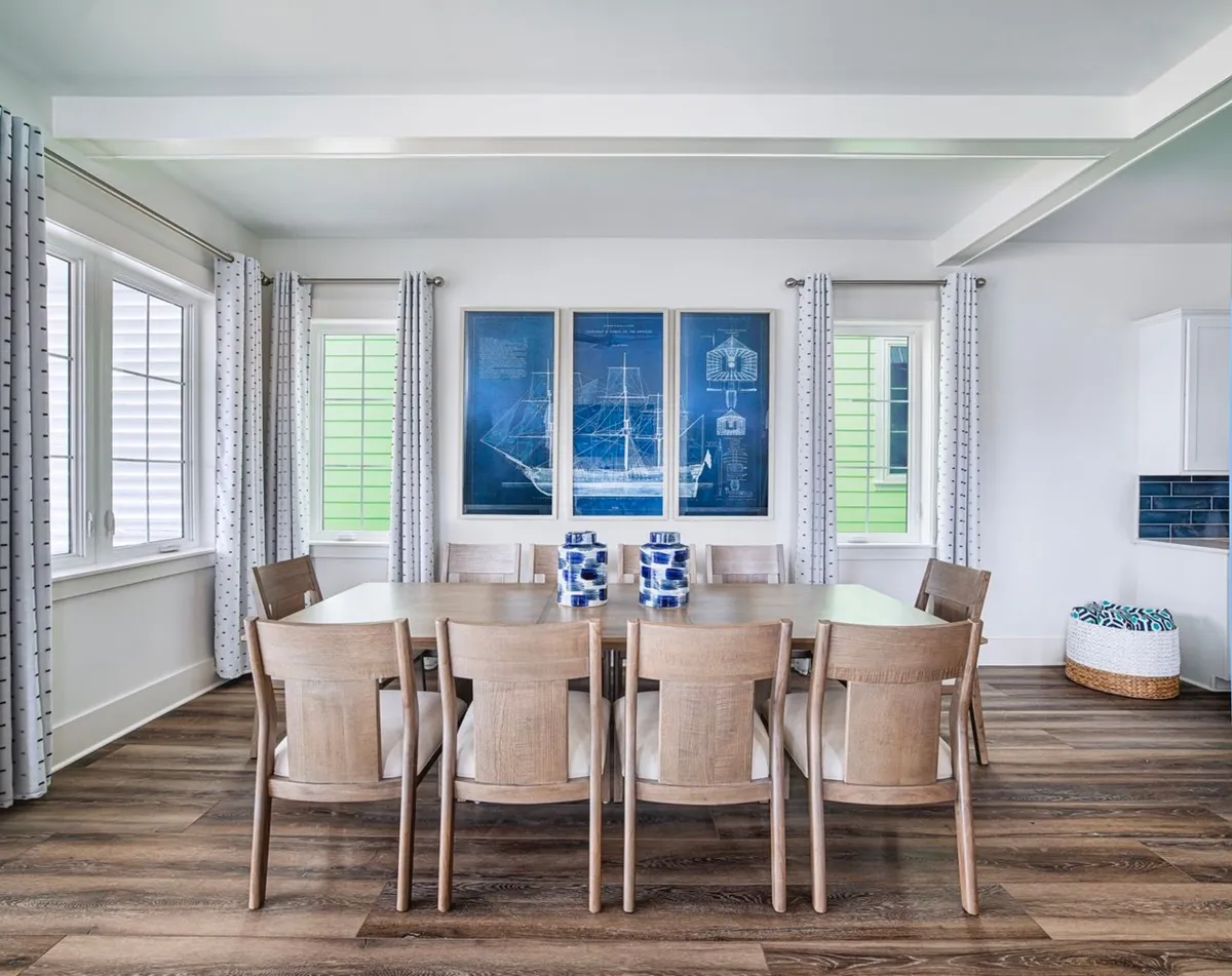 A large wooden dining table with eight chairs takes center stage, set beneath triptych nautical blueprint artwork. Sunlight pours through windows with white, patterned curtains, illuminating the space and creating a welcoming, coastal-casual mood. The wood-look flooring and coastal touches reinforce the beach retreat vibe, making this the perfect spot for group meals and lively conversation.