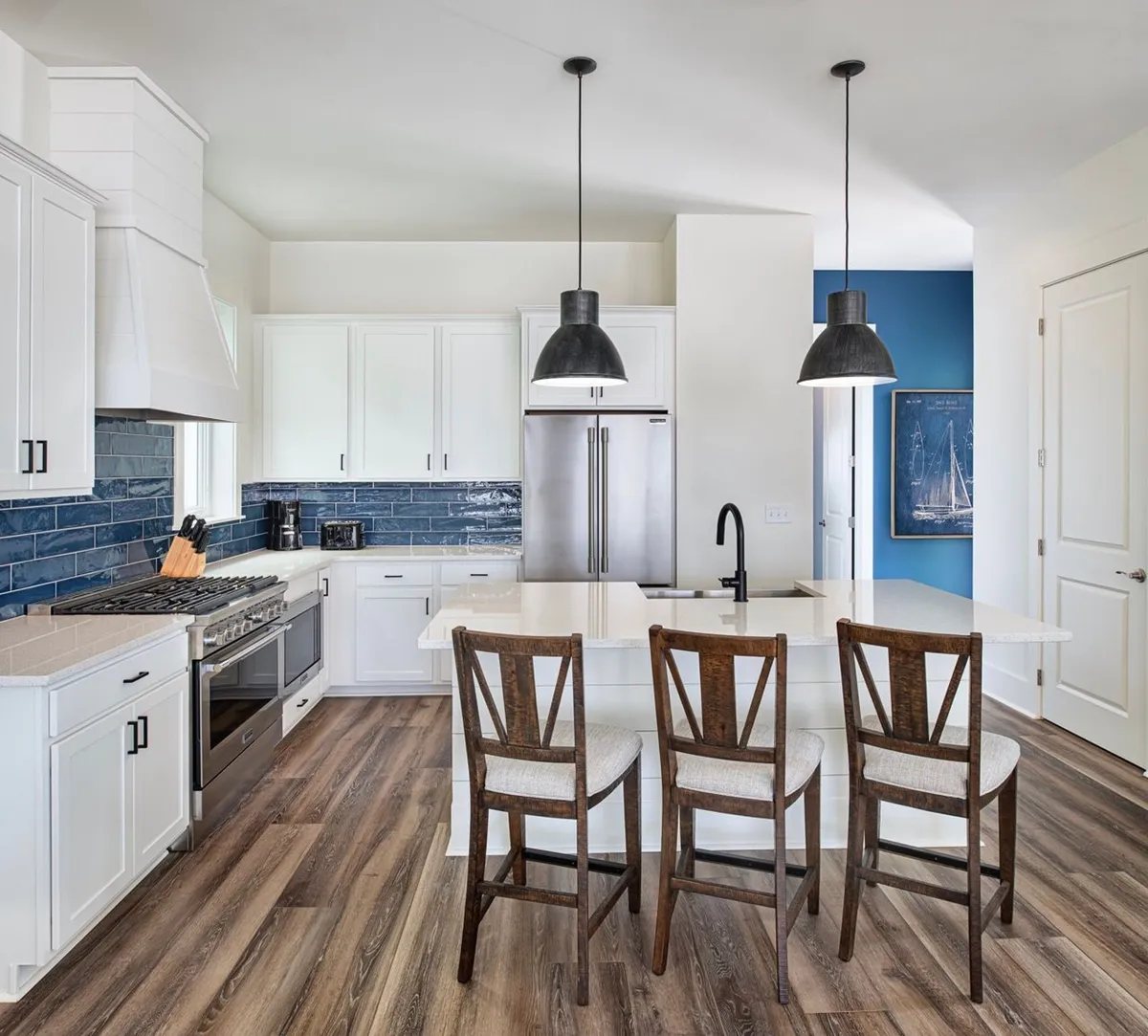 A bright, inviting kitchen features crisp white cabinetry, a striking blue tile backsplash, and a central island with seating for casual dining or cocktails. Black pendant lights add contemporary flair, while the island provides a spot for guests to gather, snack, or socialize during meal prep. The kitchen flows naturally into the rest of the living space, supporting a breezy, communal vibe.