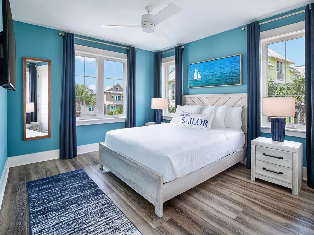 A charming bedroom with a white wooden bed, 'Sailor' pillow, large windows overlooking neighboring palm trees and coastal homes, nautical artwork, and blue curtains with matching bedside lamps. The space is bright, airy, and decorated in a soothing beach cottage style.