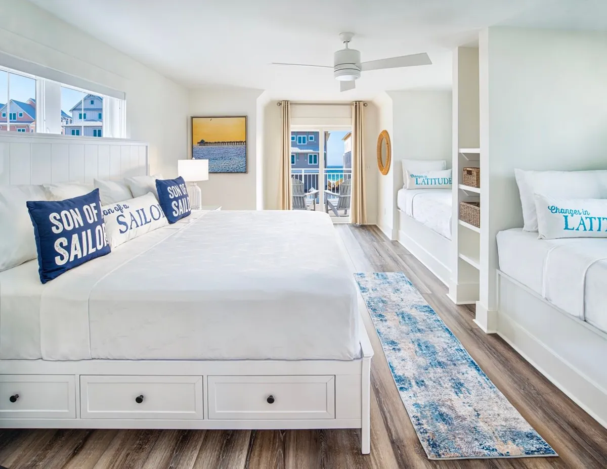This image features a large white bed adorned with navy blue 'Son of a Sailor' pillows, built-in twin beds with coastal-themed pillows, a sliding glass door leading out to a balcony with sweeping ocean views, and coastal artwork on the wall. The room is filled with natural light and evokes a laid-back, beach vacation atmosphere.