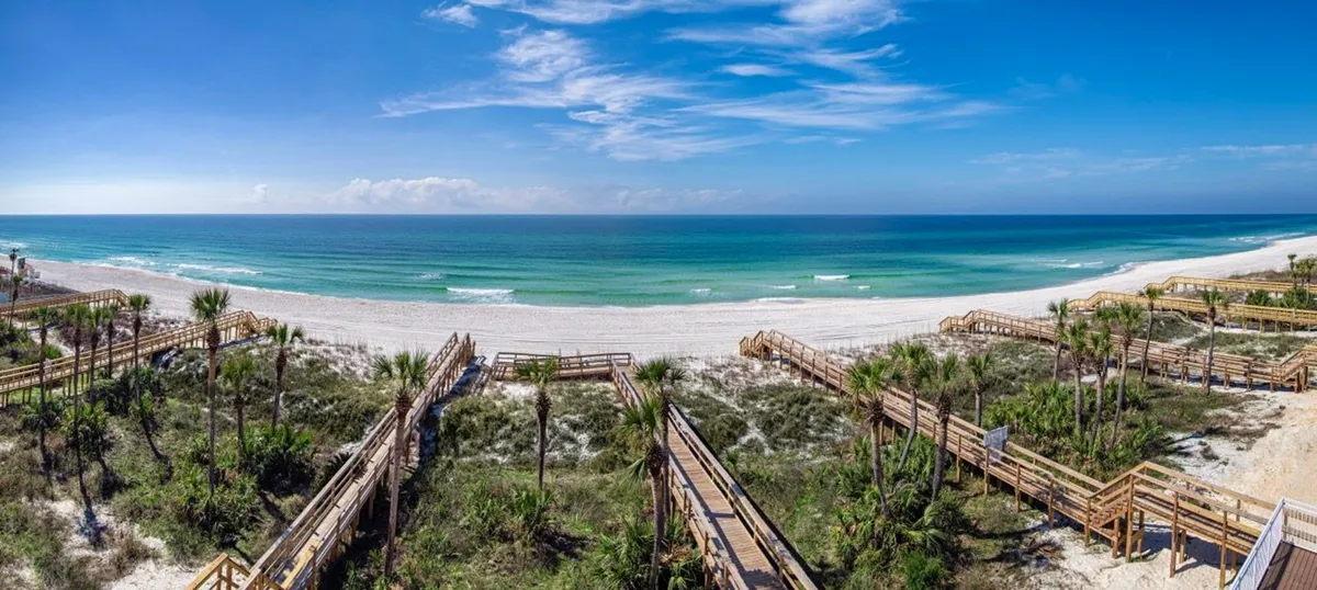 This image features wooden walkways leading directly to a pristine, white sandy private beach and clear turquoise ocean under a blue sky with scattered clouds. The scene radiates tranquility and invites families to begin their adventures right at the water’s edge.