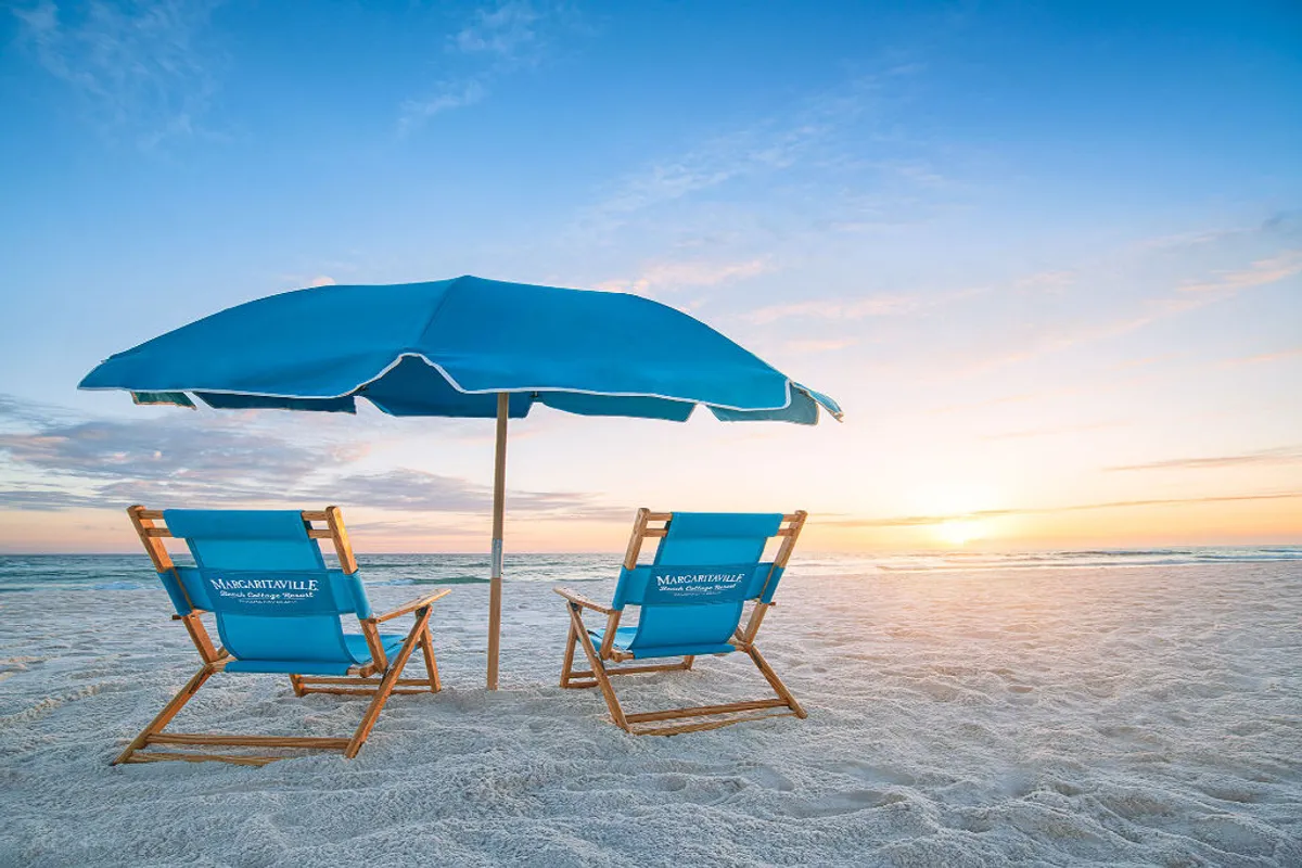 Two branded blue beach chairs and a large umbrella are positioned on a wide expanse of pristine sand, facing the setting sun over the Gulf of Mexico. The atmosphere is serene, inviting, and distinctly beach resort, evoking relaxation and the allure of private beach access.