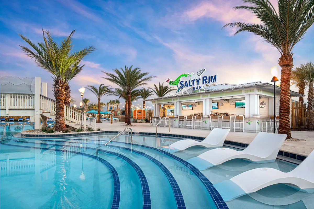 The Salty Rim Bar with its illuminated signage overlooks a curved swimming pool with submerged lounge chairs, surrounded by tall palm trees and inviting seating areas. The scene is set for relaxed poolside dining and sunset cocktails.