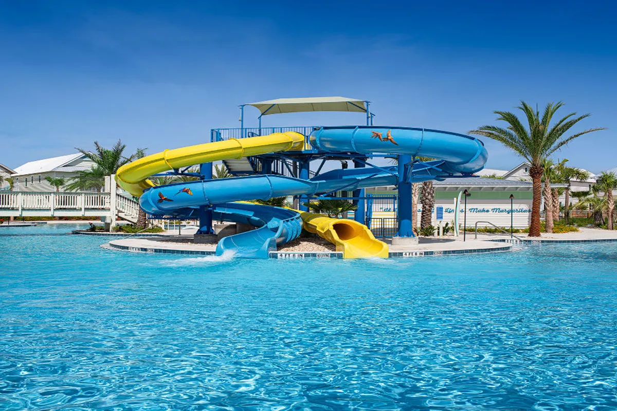 A vibrant, energetic scene highlights colorful yellow and blue water slides curving into a large resort pool surrounded by palm trees and a clear blue sky. Families and children are seen enjoying waterslide thrills and swimming in the pool.
