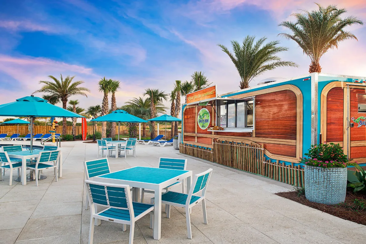 Featuring the poolside Salty Rim Bar & Grill with its colorful signage and wooden accents, this image shows guests relaxing at shaded tables under blue and white umbrellas. Palm trees line the area, and sun loungers are visible in the background, emphasizing the availability of both lively and laid-back hangouts at the resort’s heart.