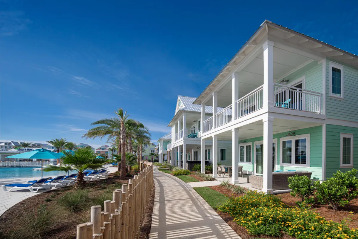 This image captures the exterior of a light turquoise coastal-style house with expansive balconies overlooking a vibrant pool area. Blue umbrellas and lounge chairs invite relaxation, while palm trees and lush tropical landscaping frame the scene. A wooden post fence lines the sidewalk, and the setting exudes an inviting, leisurely holiday atmosphere.